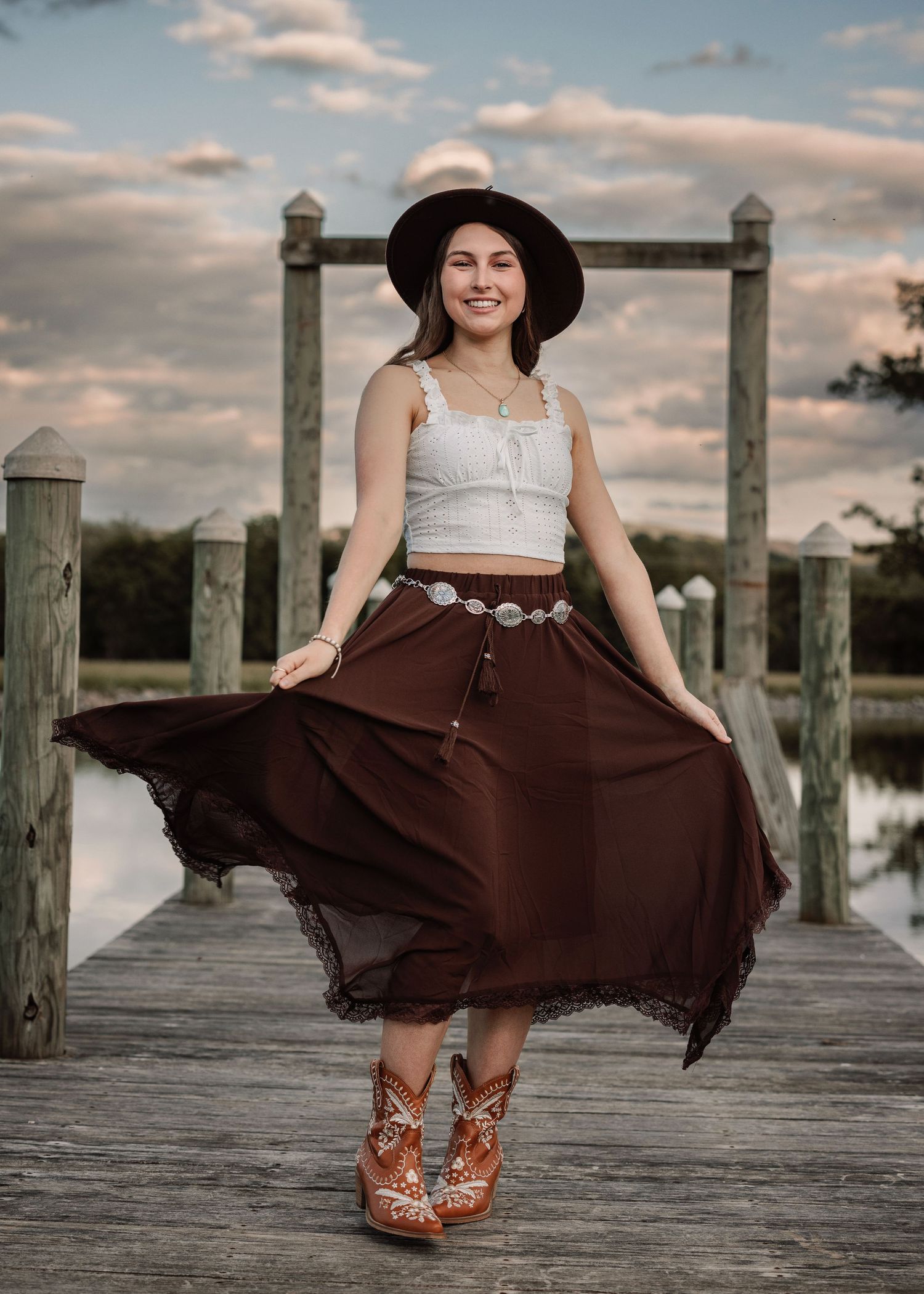 Person wearing flowing brown skirt and hat twirls on wooden pier during golden hour.
