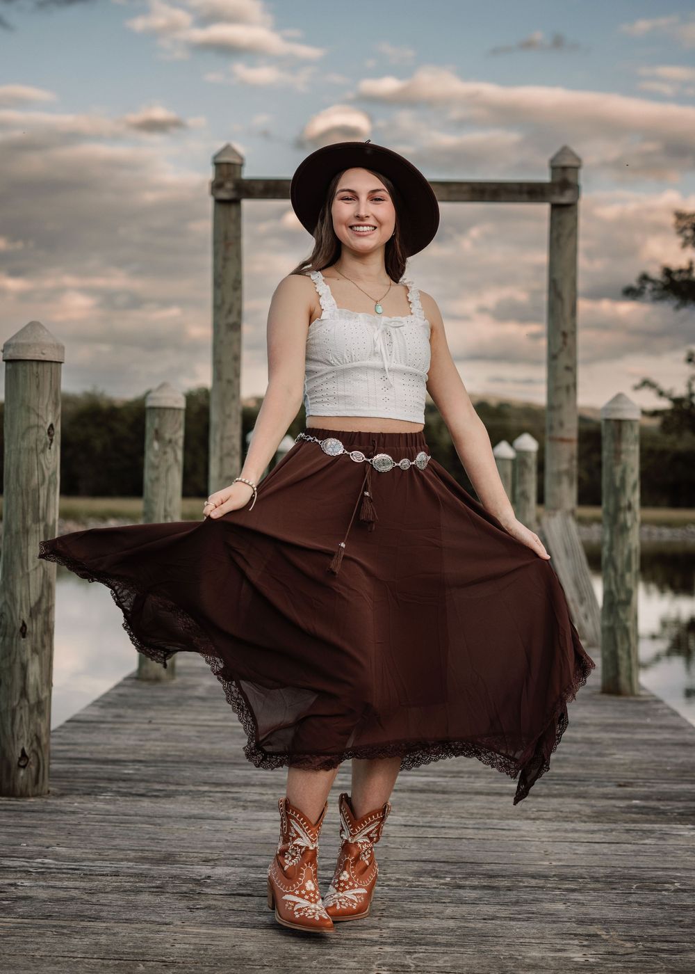 Person wearing flowing brown skirt and hat twirls on wooden pier during golden hour.