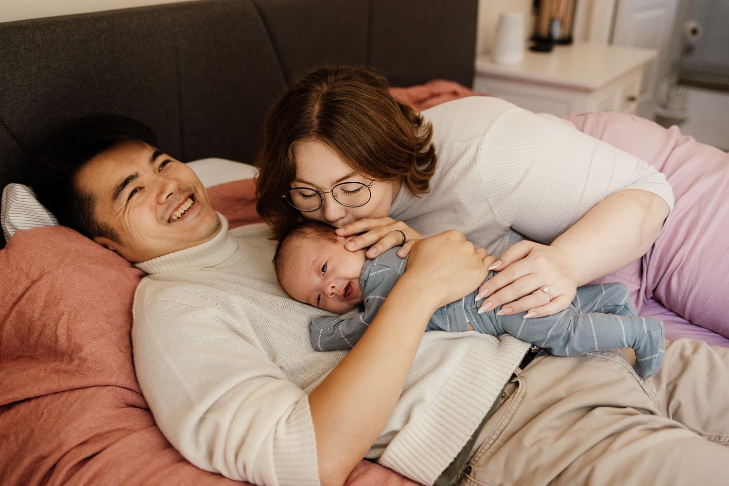 Mom and dad laying on a bed with their newborn - in-home family session Bay Area