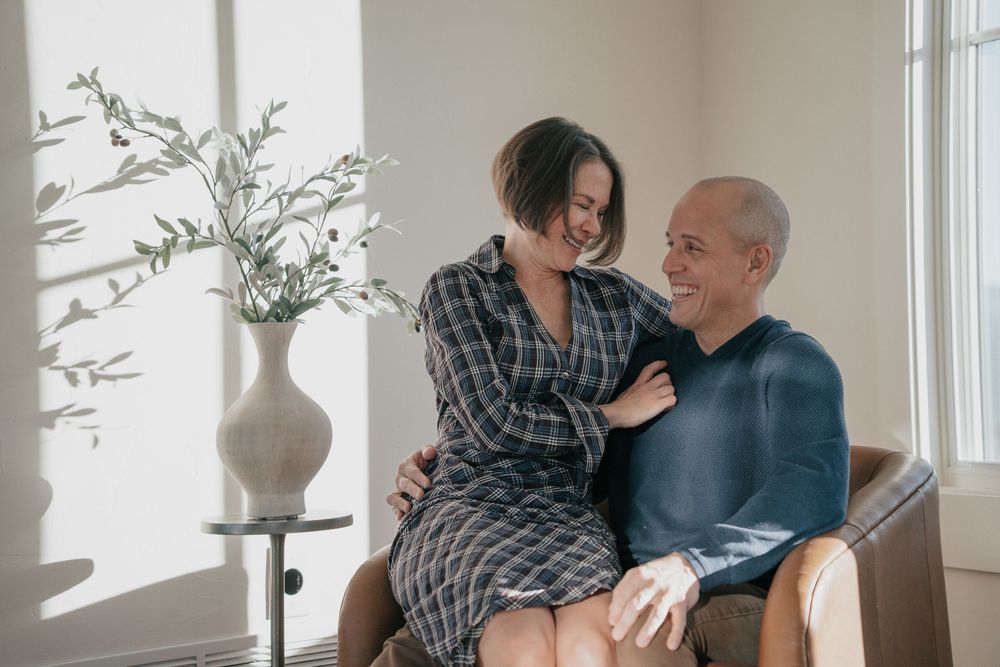 Two people sitting together in bright natural lighting with white curtained windows and potted plants. Colorado photographer couples natural