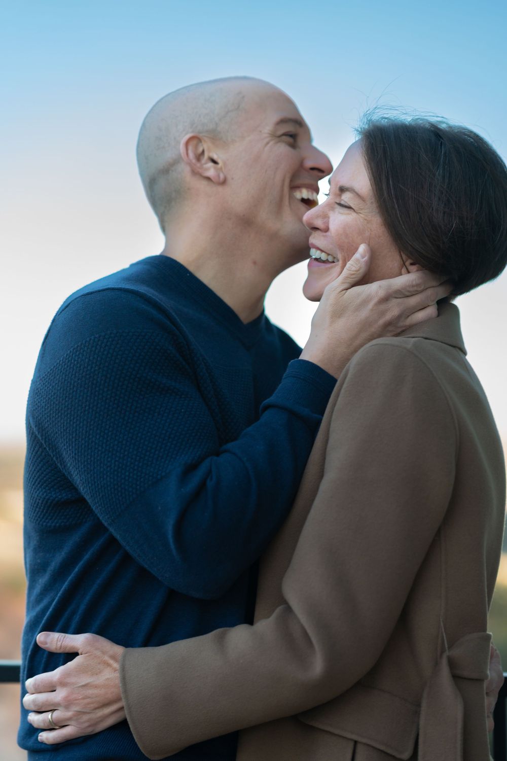 A loving couple shares an intimate moment of laughter and joy against a soft blue sky background. Outdoor couples session Colorado Mantiou