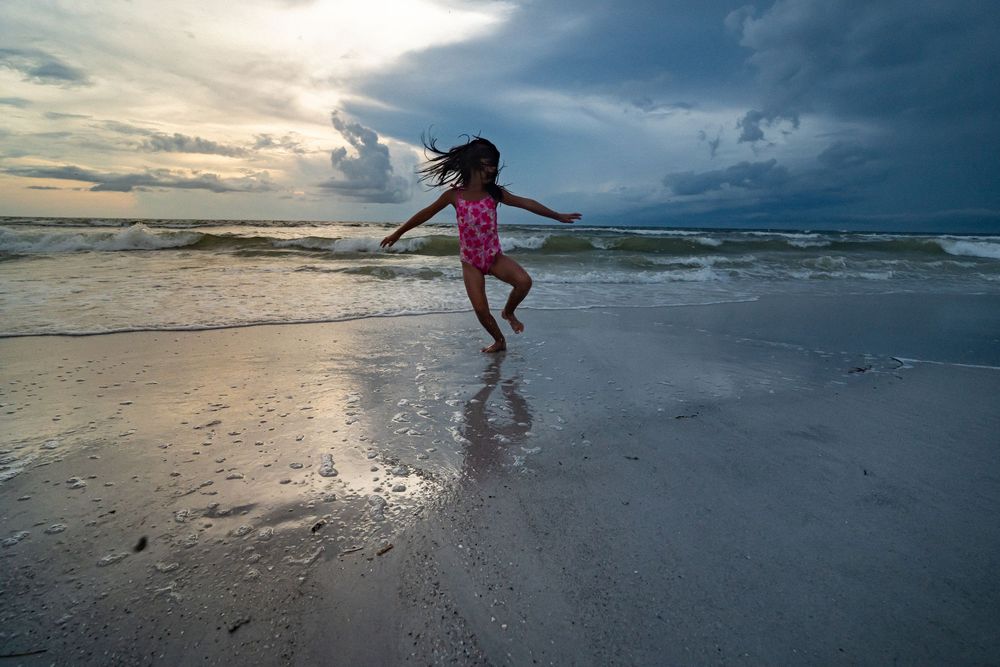 Silhouetted figures play and jump on a misty beach at sunset with dramatic cloudy skies above. Florida family photographer
