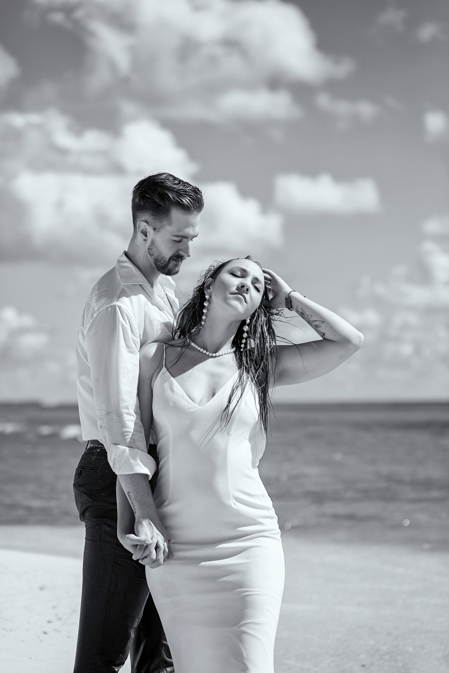 A romantic black and white beach portrait of a couple embracing along the ocean shore with white clouds above.