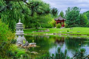 A peaceful Japanese garden features a stone lantern and red pagoda pavilion beside a tranquil pond surrounded by willow trees.