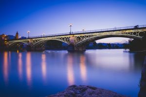 A stone bridge spans across calm blue waters at twilight with illuminated arches reflecting in the smooth river surface.