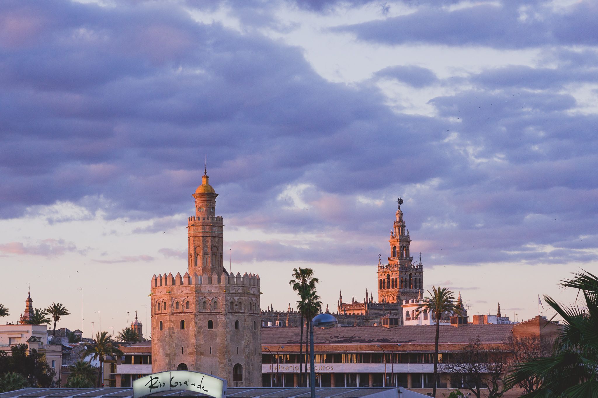 Historic cathedral spires and towers stand against a dramatic cloudy sky in Seville, Spain at sunset.