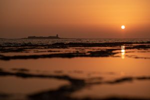 Stunning orange sunset over calm ocean waters with silhouetted lighthouse and reflective waves at dusk.