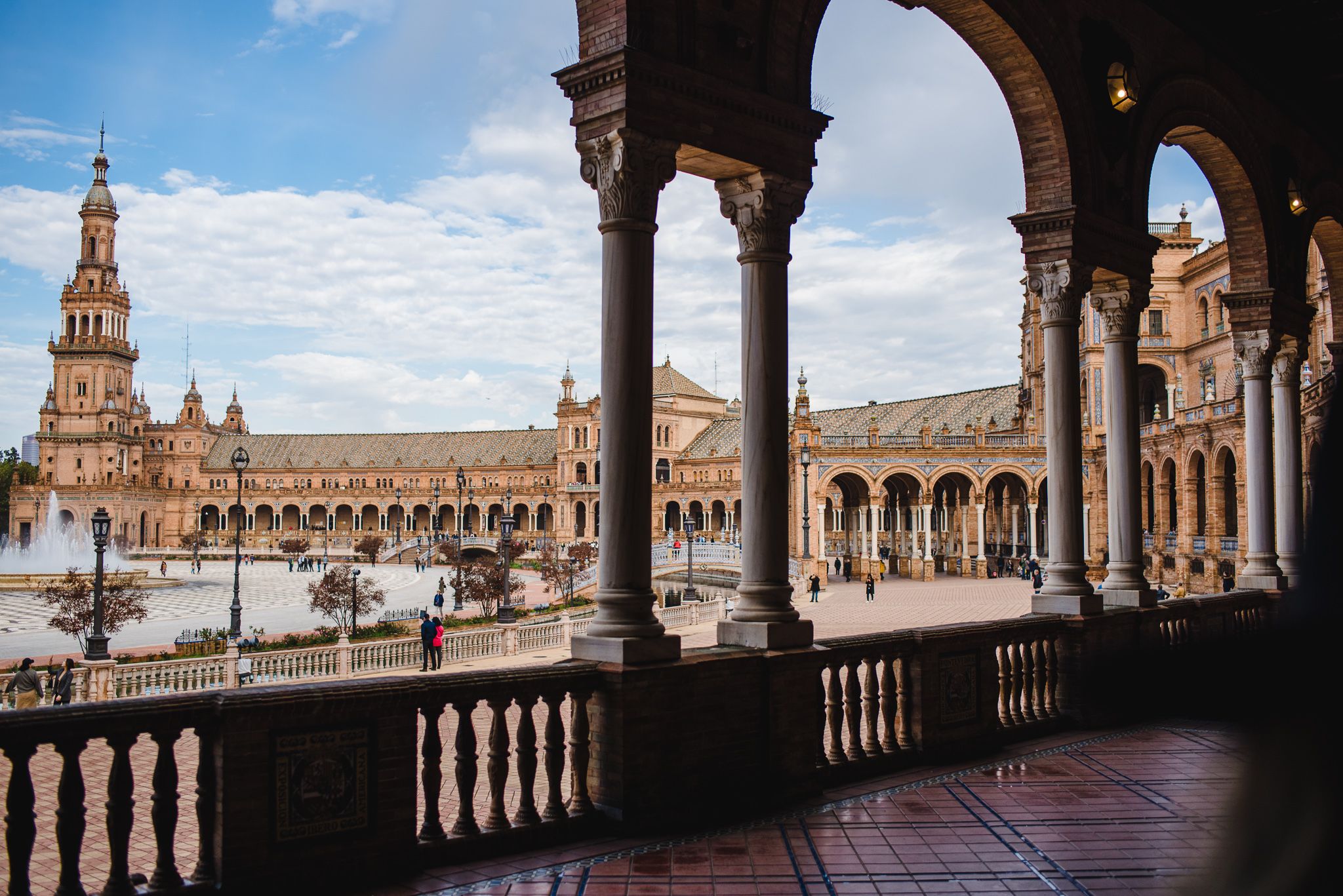 View of Plaza de España in Seville through ornate arches, featuring Renaissance architecture and iconic tower under blue sky.