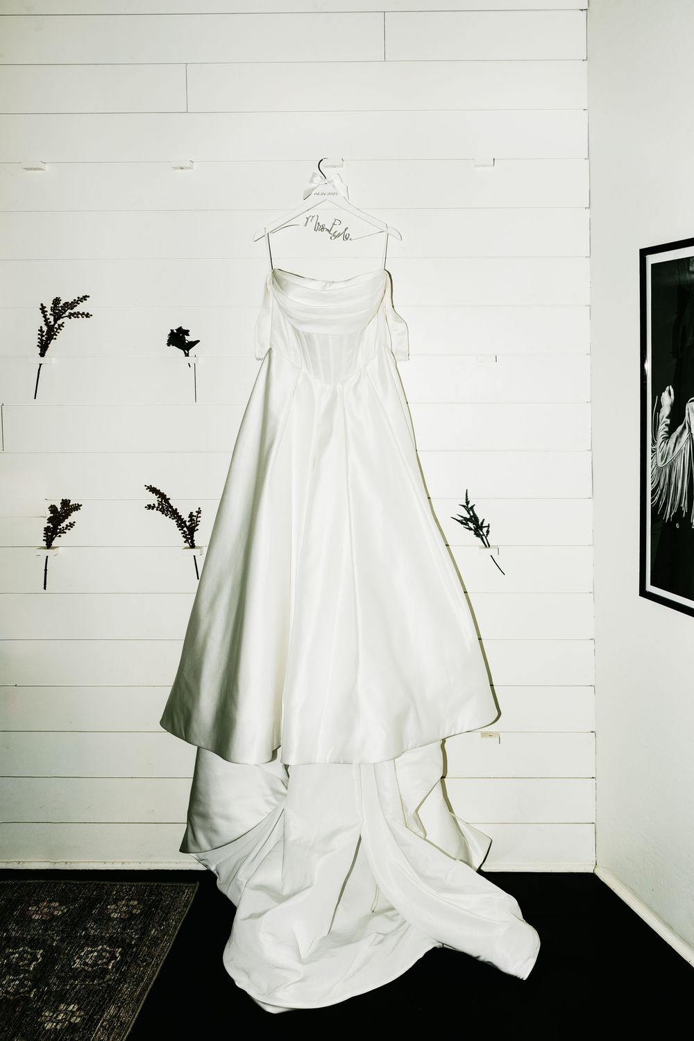 White wedding dress hangs elegantly against white paneled wall in minimalist setting.