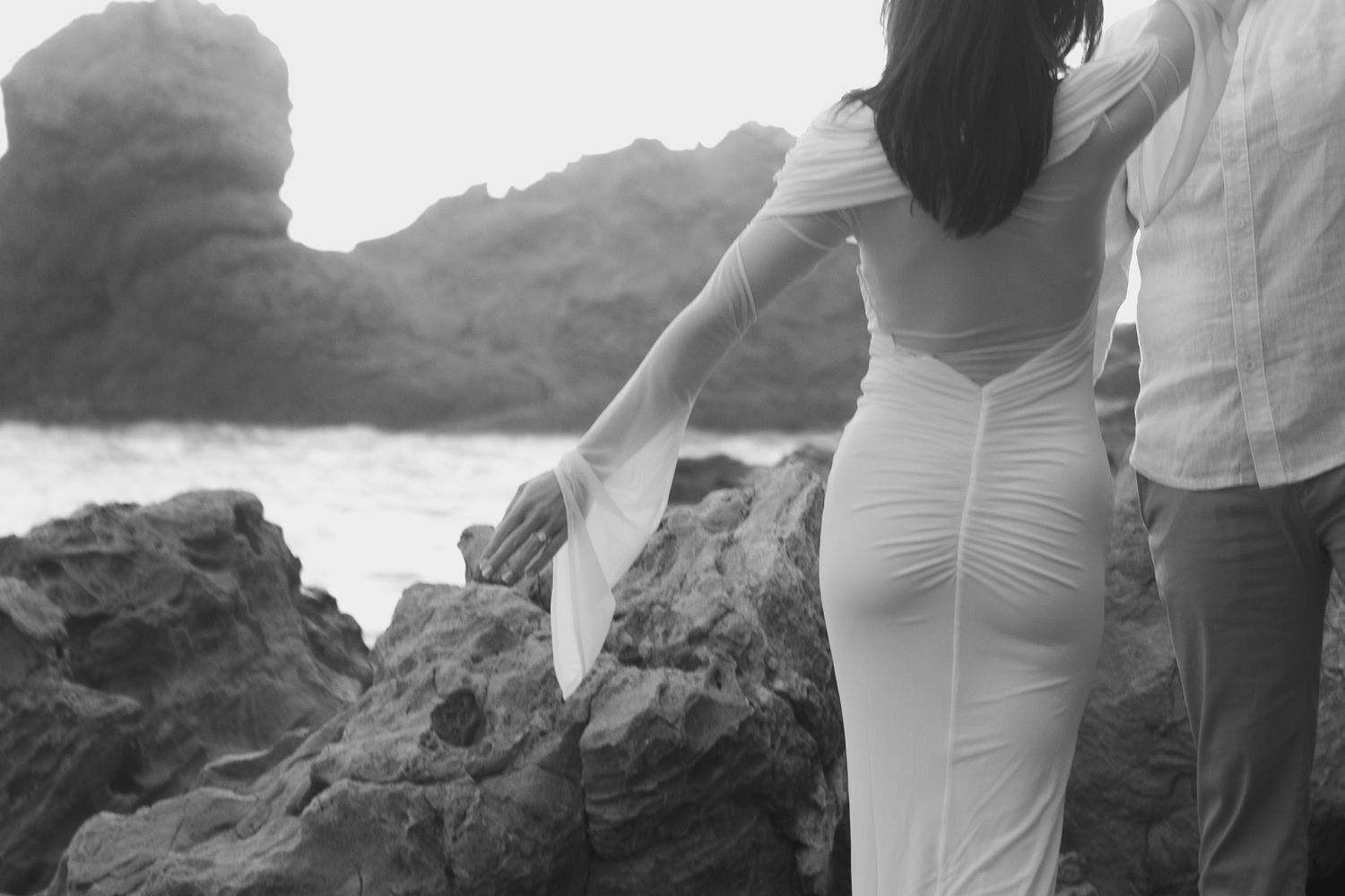 Black and white photo of a couple in formal attire standing on coastal rocks near the ocean.