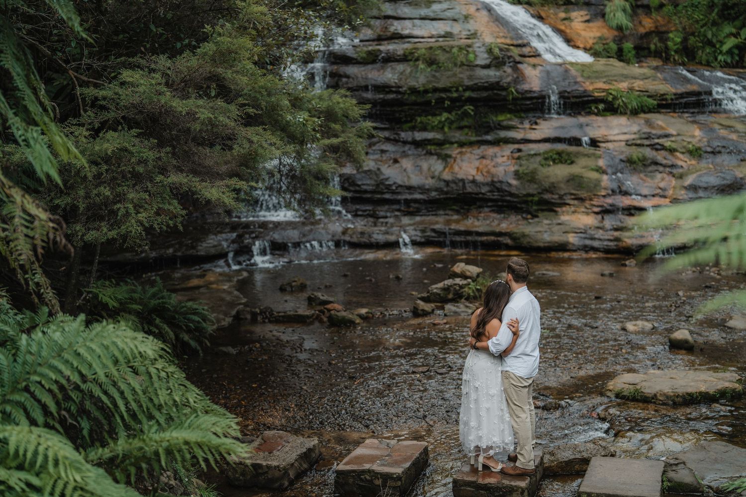 A couple embraces near a cascading waterfall in Blue Mountains surrounded by ferns and rocky cliffs.