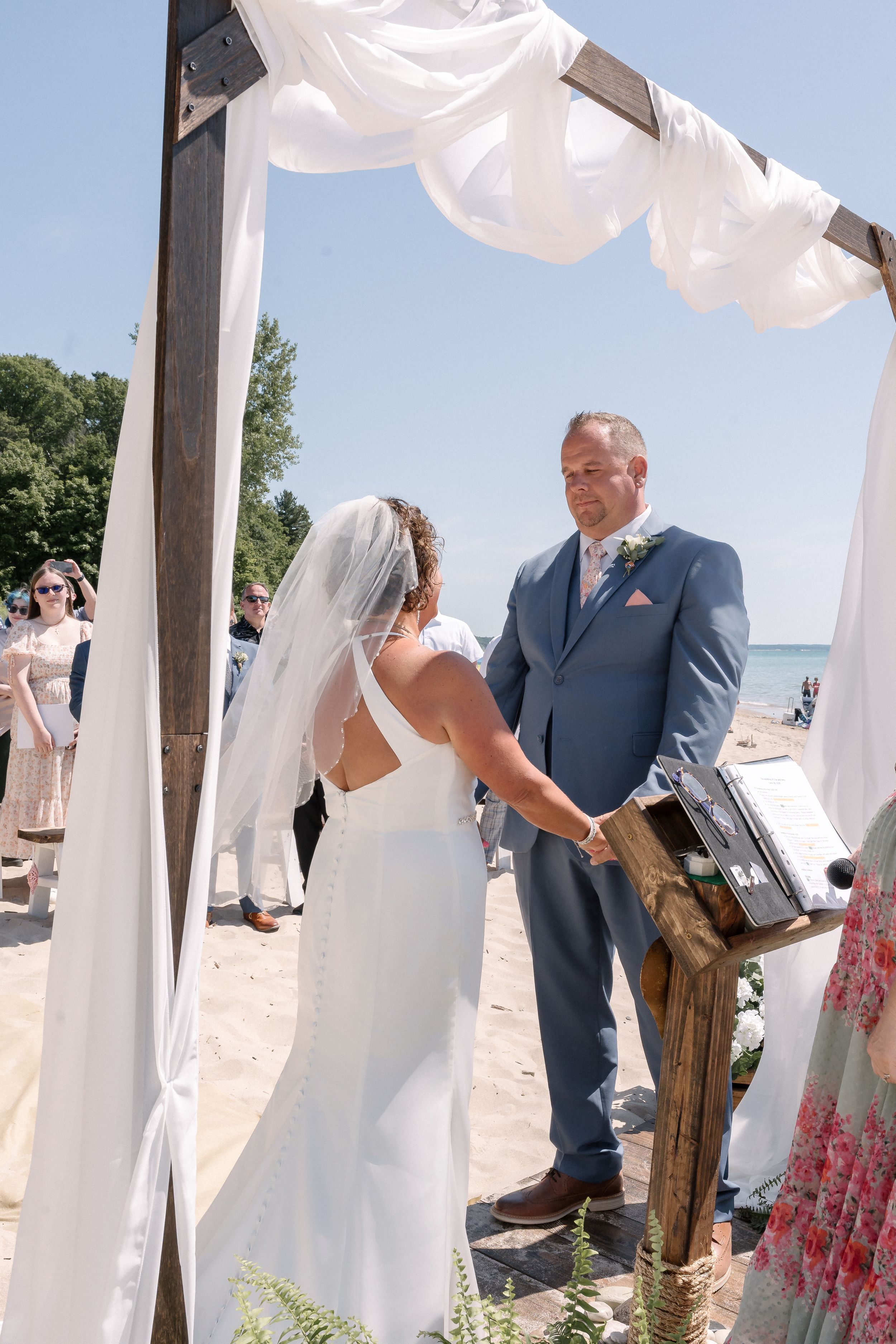 A beach wedding ceremony on a sunny day with white flowing fabric draped between wooden posts by the ocean.