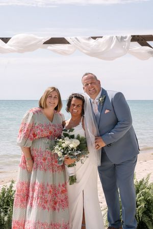 A beachside wedding ceremony with three people posing together under flowing white fabric by the ocean.