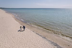 Two figures walk along a tranquil shoreline beside calm blue waters on a sunny day.
