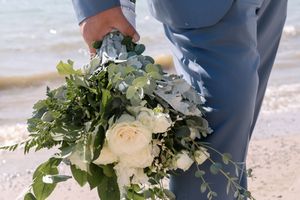 A hand wearing a blue suit sleeve holds a delicate white and green floral bouquet against a beach backdrop.