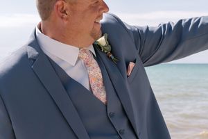 A groom dressed in a gray suit with a peach tie smiles at a beach wedding ceremony.