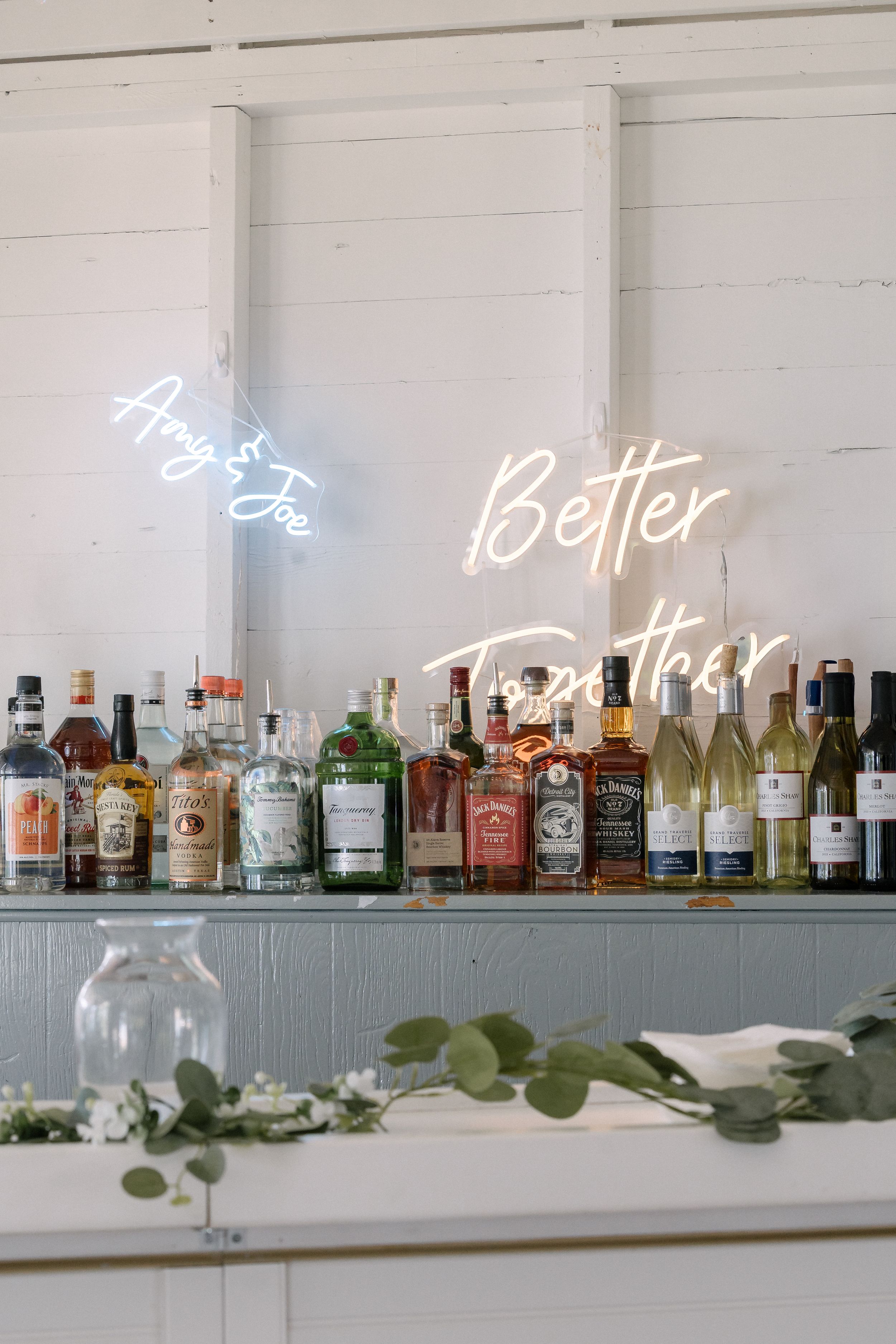 White bar setup with neon signs reading 'Better Together' displayed above lined up liquor bottles and greenery decoration.
