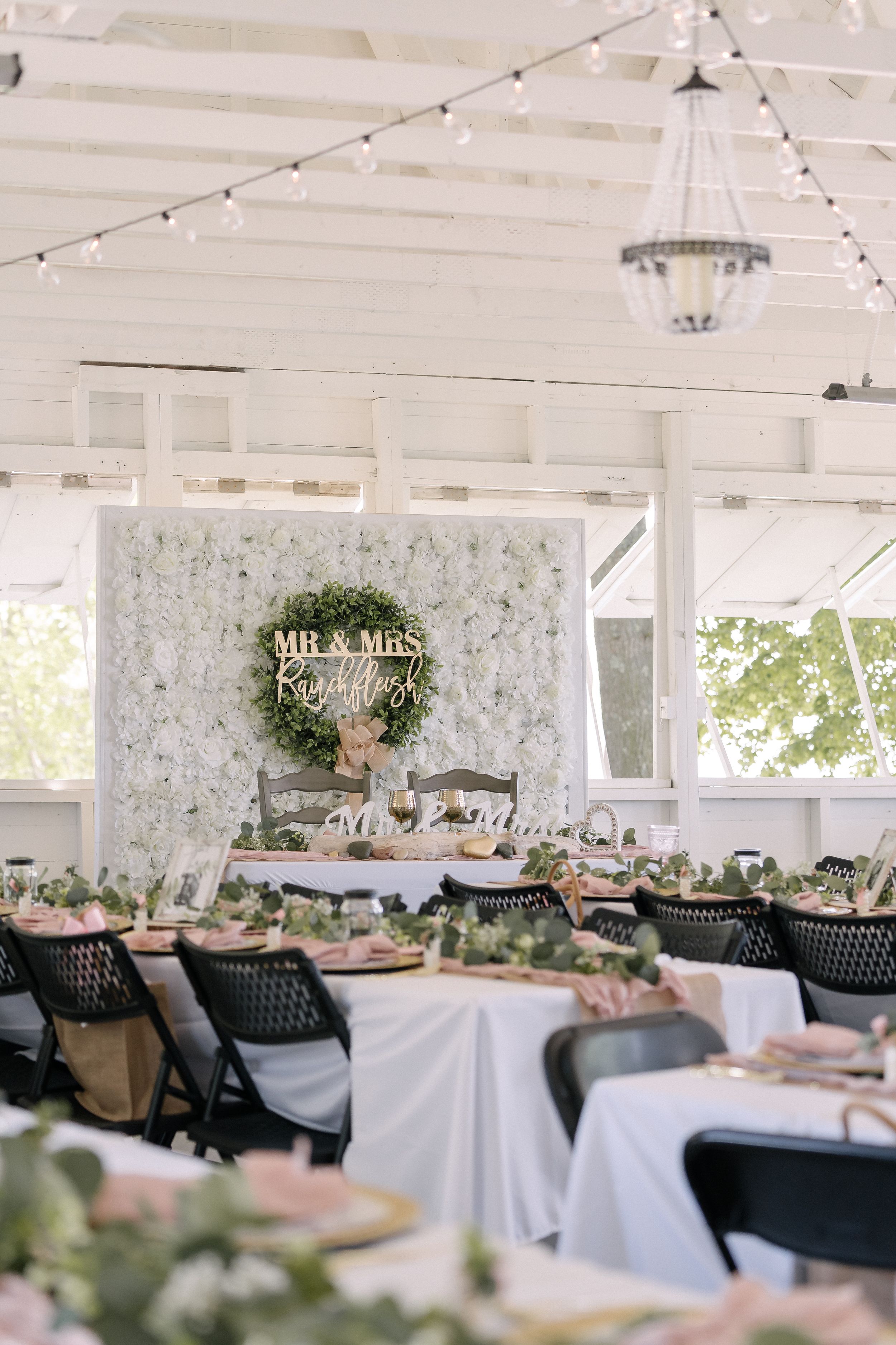 Elegant wedding reception setup with white floral backdrop, Mr & Mrs sign, and black chairs under a covered outdoor pavilion.