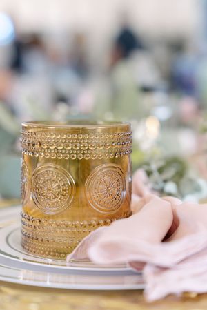 Ornate amber glass votive holder with embossed medallion pattern sits on plate beside blush pink napkin at elegant place setting.