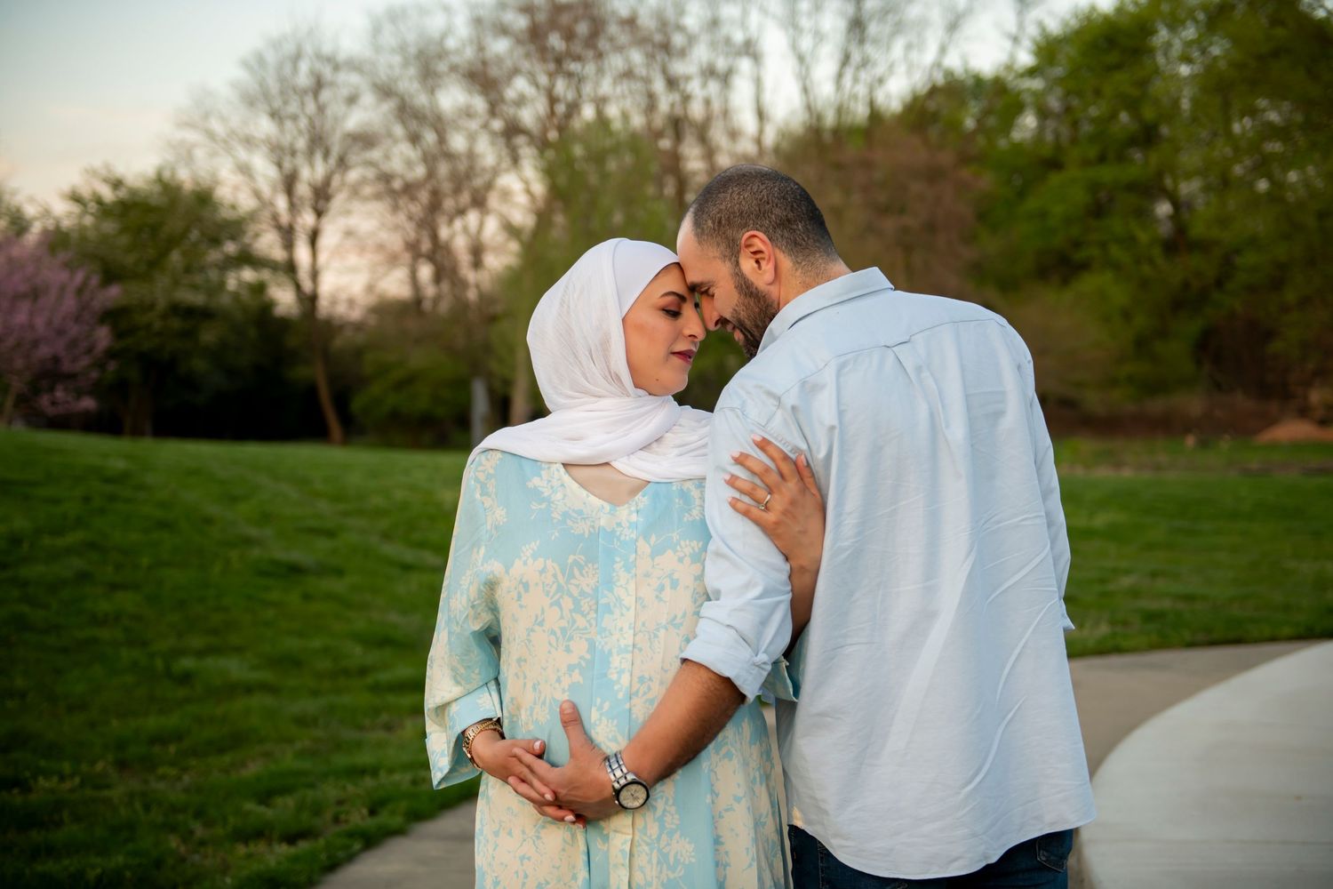 Couple embraces tenderly during sunset photo session on park path with green spring foliage in background.
