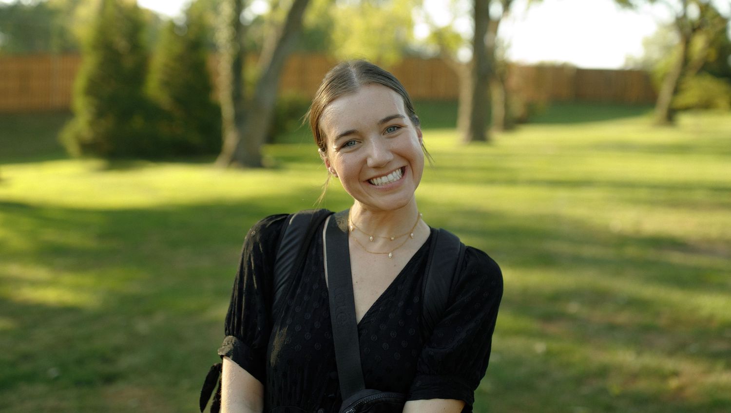 Person wearing a black top smiles warmly while standing in a sunny park with green trees in the background.