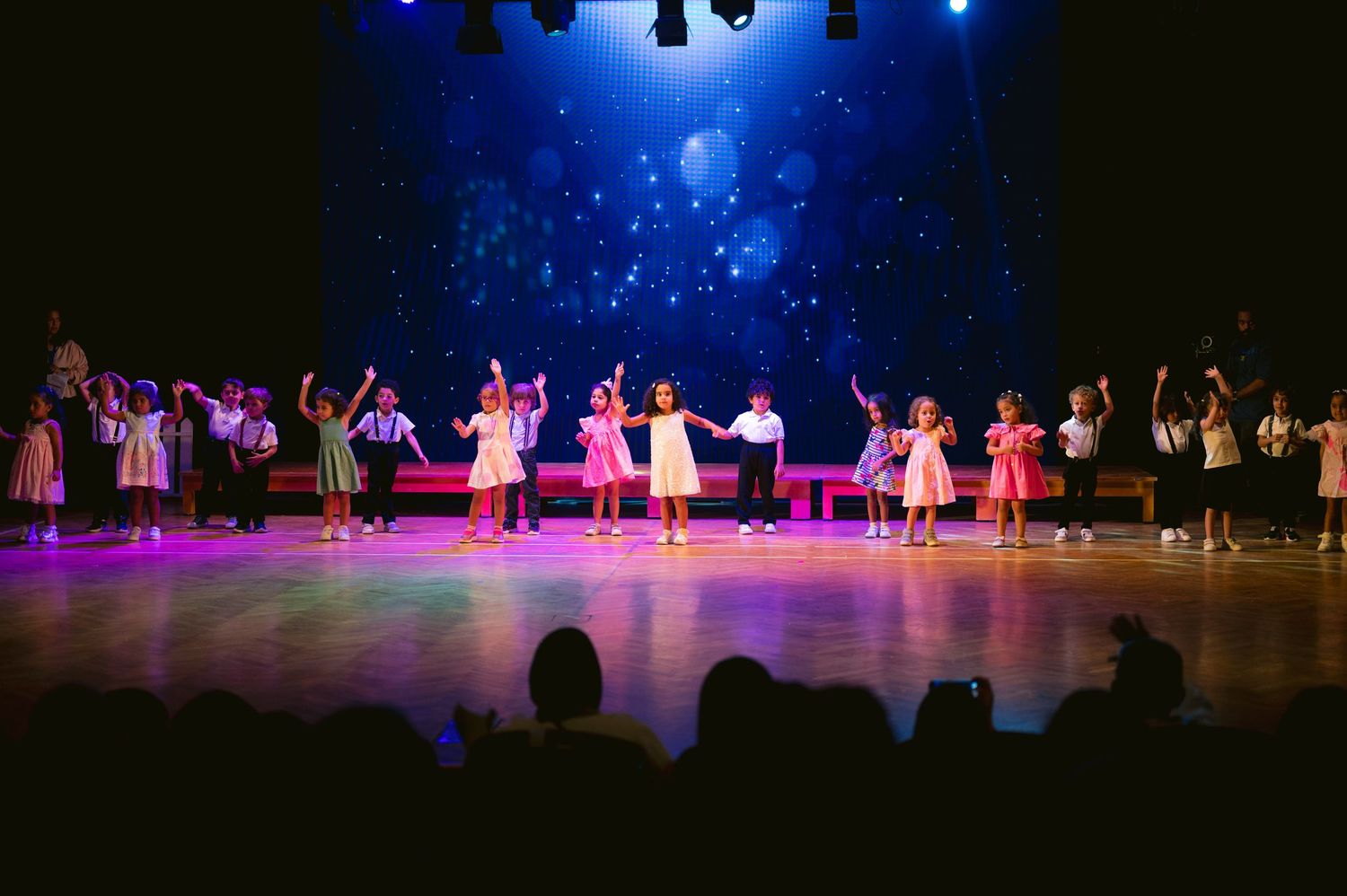 Performers in colorful dresses dance on a dimly lit stage with blue spotlights and silhouetted audience members visible.