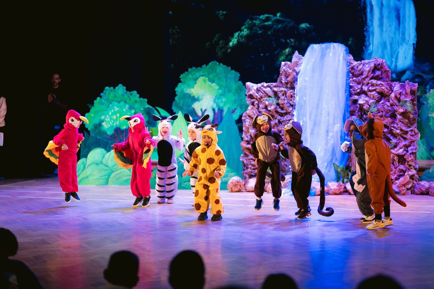 Costumed performers dance in formation on a colorfully lit stage with theatrical scenery.