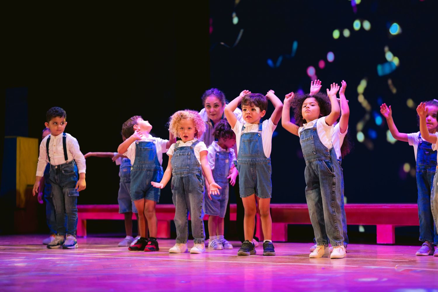 Children perform together on stage in matching outfits under colorful theatrical lighting during a dance recital.