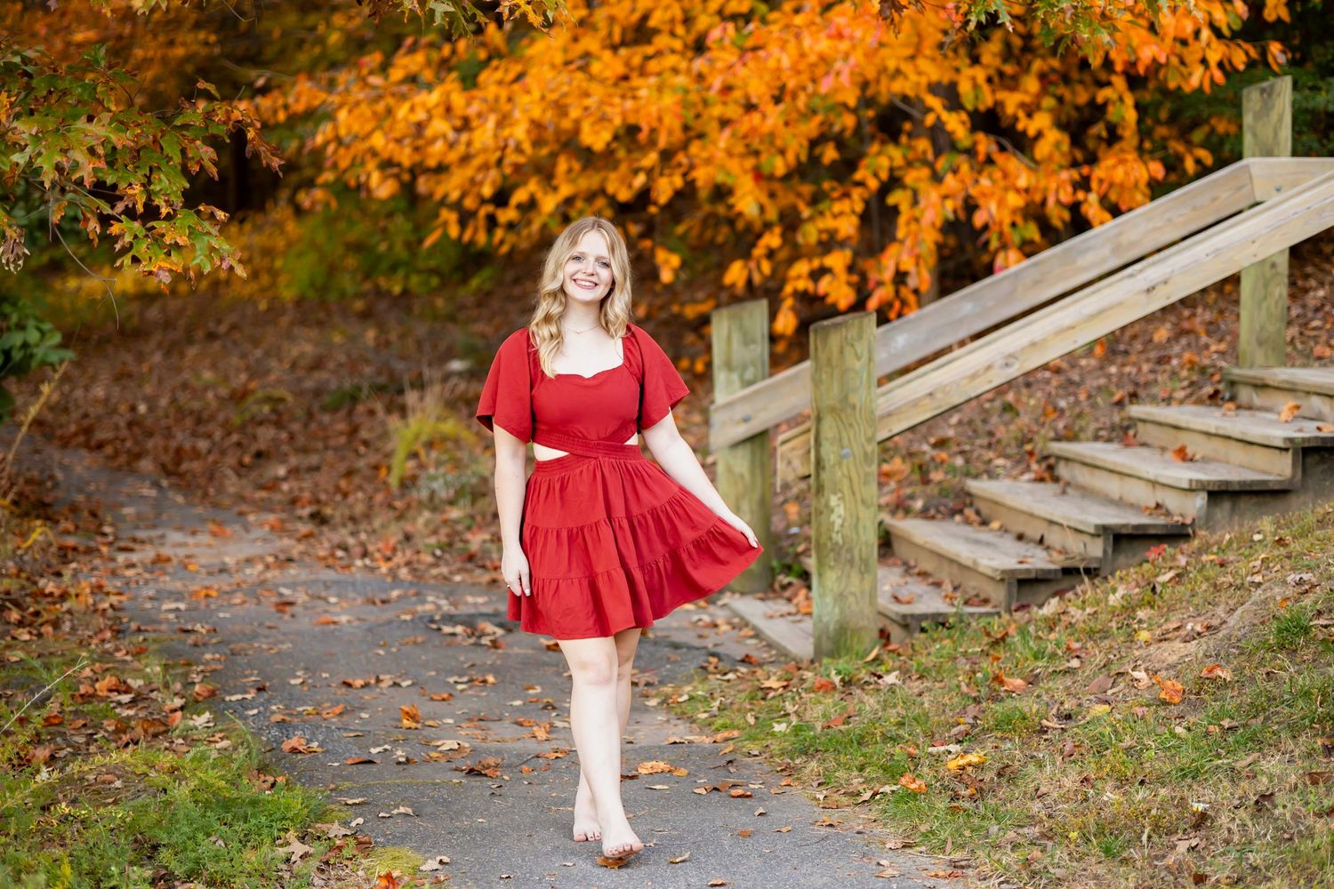 Person in a flowing red dress twirls among vibrant orange autumn foliage and wooden steps in an outdoor setting. fall senior photo session