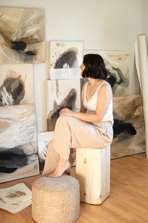 Abstract paintings decorate the walls of a bright room with a figure seated on a textured ottoman.