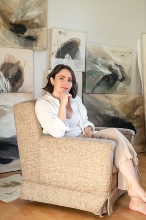 Someone sits on a beige upholstered chair in an art gallery space with abstract paintings.