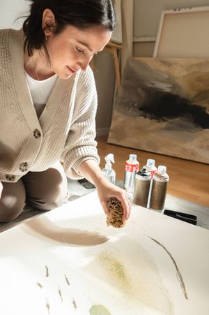 Artist sprinkles materials onto a white canvas with paint bottles and an abstract artwork visible in background.