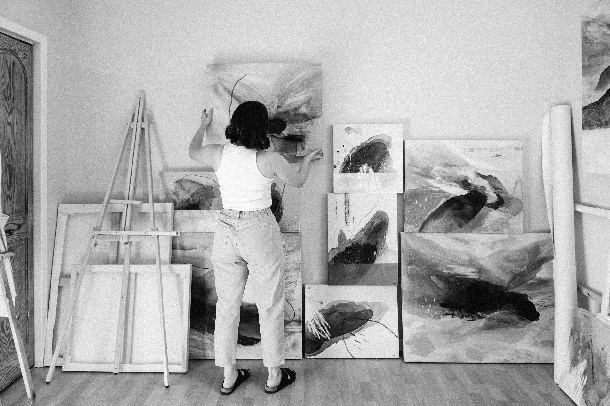 A series of black and white photographs show canvases and artworks arranged against walls in a gallery-like setting.