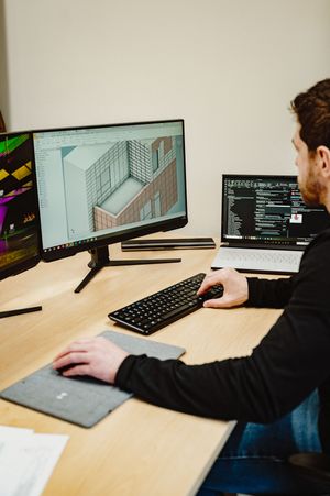 Designer working at desk with 3D architectural render on monitor and coding IDE on second screen.