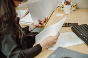 Student taking notes with pencil while studying at desk with laptop and documents.