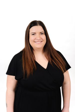 Professional studio portrait of a smiling person with long brown hair wearing a black v-neck top against white background.
