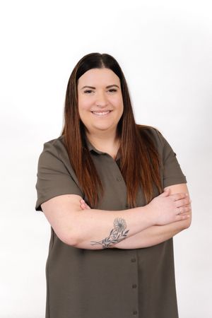 Professional studio portrait showing someone in an olive green button-up shirt with visible arm tattoos against white backdrop.