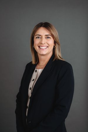 Professional woman in black blazer smiling confidently against gray background for corporate headshot.