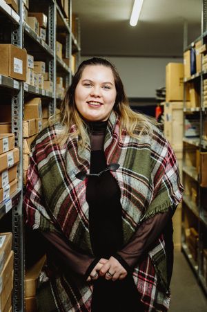 Person wearing a plaid shawl stands smiling in a warehouse storage area with shelves of boxes behind them.