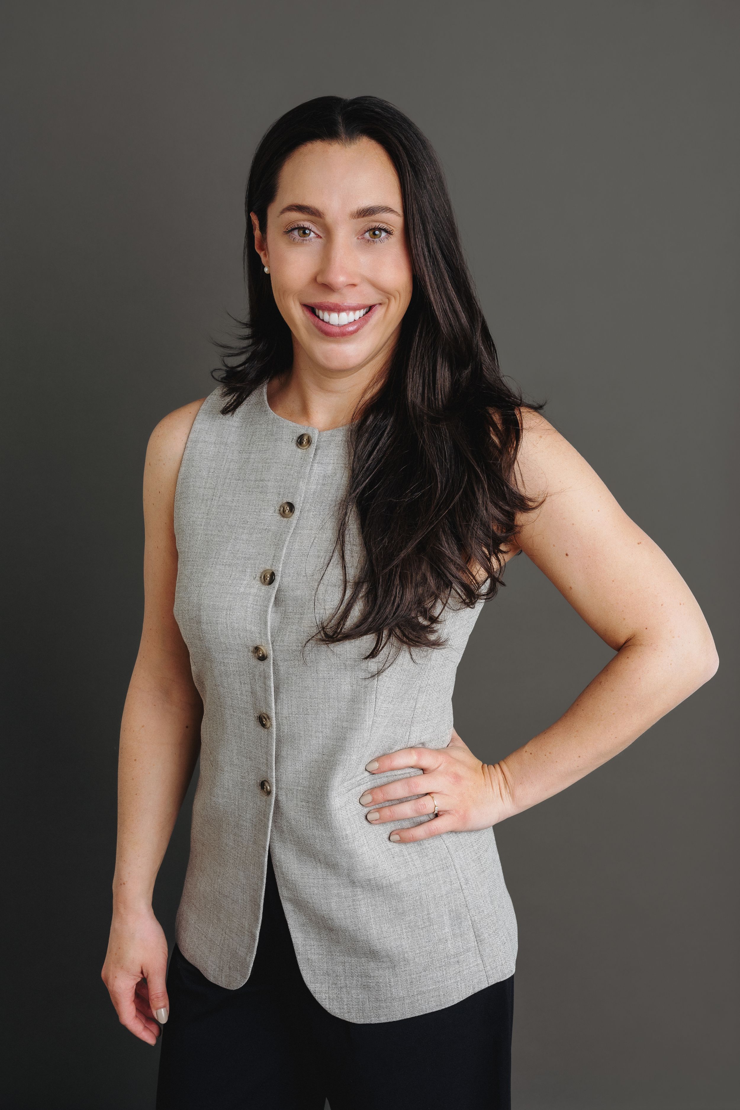 Professional woman in gray sleeveless blazer smiling confidently against dark background.