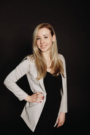 Professional portrait of a woman in a white blazer and black top posing against a dark background.