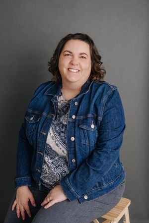 Smiling woman in denim jacket and patterned top seated on stool against gray background.