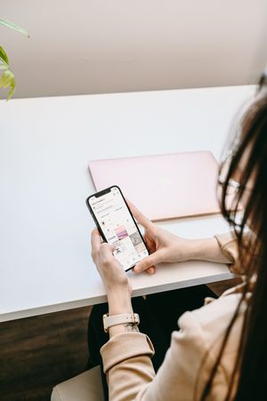 Person holding and scrolling through a smartphone while wearing a rose gold watch and sitting at a wooden desk.