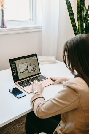 Working remotely at a minimalist white desk setup with a laptop, phone and indoor plant by window.