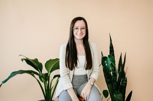 Someone sitting on a stool wearing a cream blazer and gray pants with houseplants on a beige background.