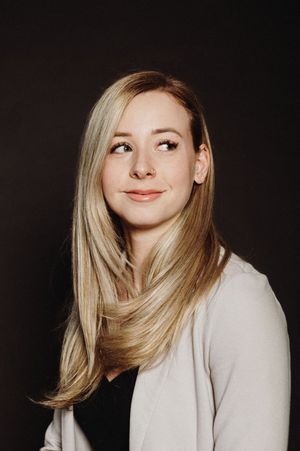 Professional portrait photo of a person with long blonde hair wearing a light cardigan over a dark top against a dark background.