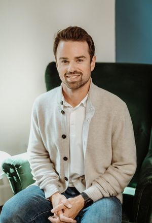 Professional portrait of someone wearing a beige cardigan and blue jeans while seated on a leather sofa.