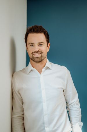 Professional headshot of a person in a crisp white button-down shirt smiling against a teal background.