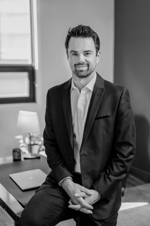 Professional black and white office portrait of a businessman in a dark suit leaning against a desk.