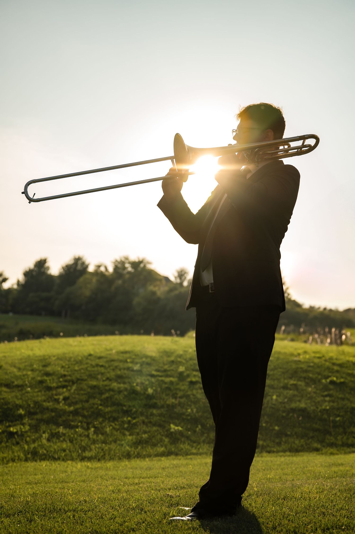 A silhouetted figure plays trombone in a grassy field at sunset with golden light shining through.
